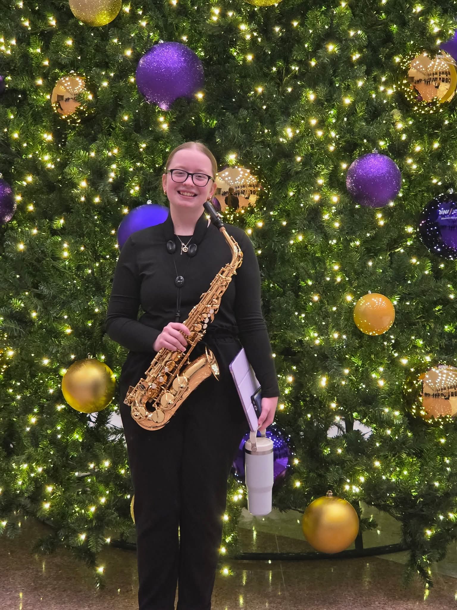 Kara McGonegle standing in front of a lighted Christmas tree, holding a saxophone.