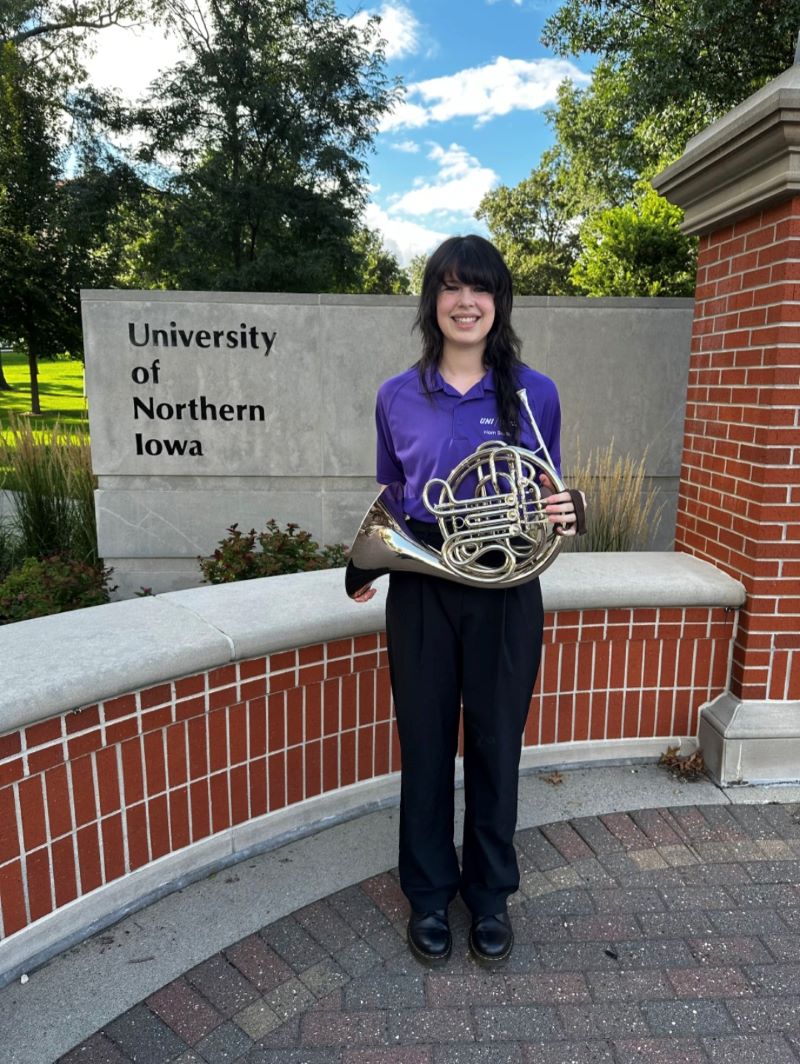 Maddie Klein standing, holding horn in front of University of Northern Iowa sign.