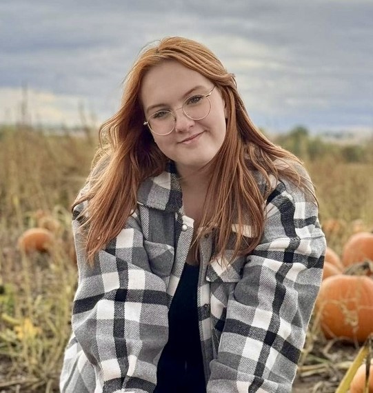 Akela Salter wearing grey and white plaid flannel, sitting in a pumpkin patch