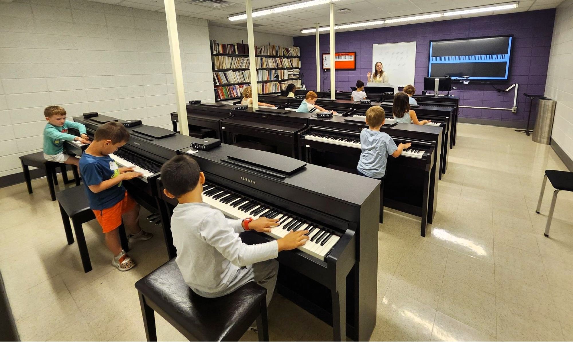 View from the back of a piano lab with students at pianos and teacher in front of room talking