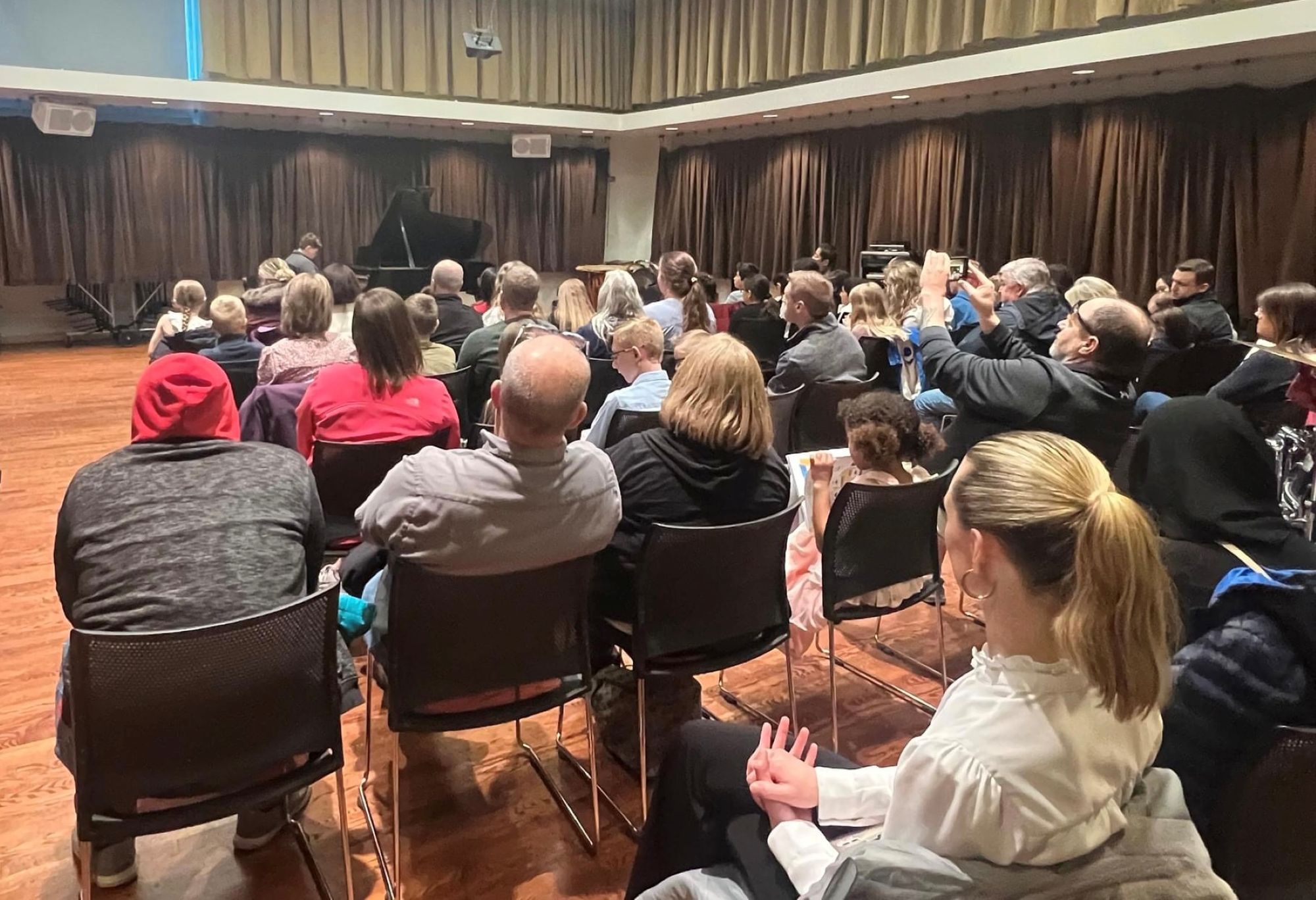 Viewing from the back of a recital. Backs of audience members in chairs with boy playing a grand piano at the front.