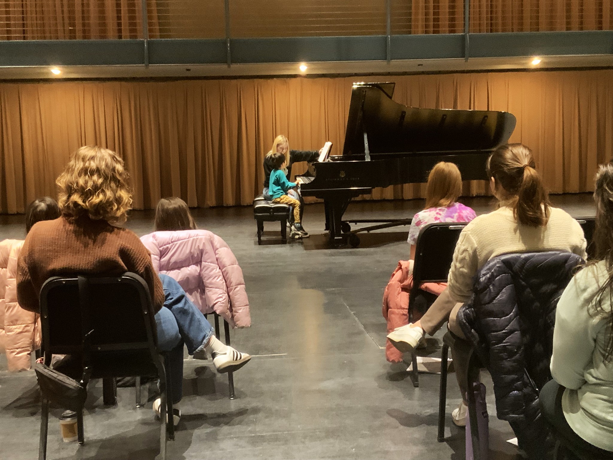 Student and teacher at the piano in front of an audience during a performance class. The teacher is pointing to something on the student's music.
