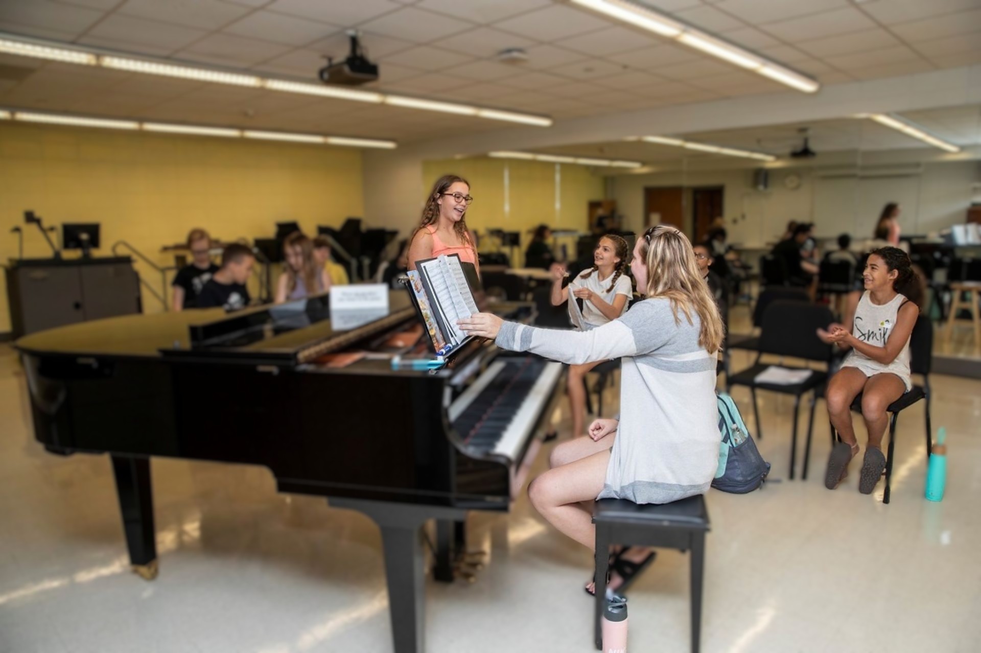 Teacher sitting at grand piano looking at female student that is standing by the piano singing with friends applauding