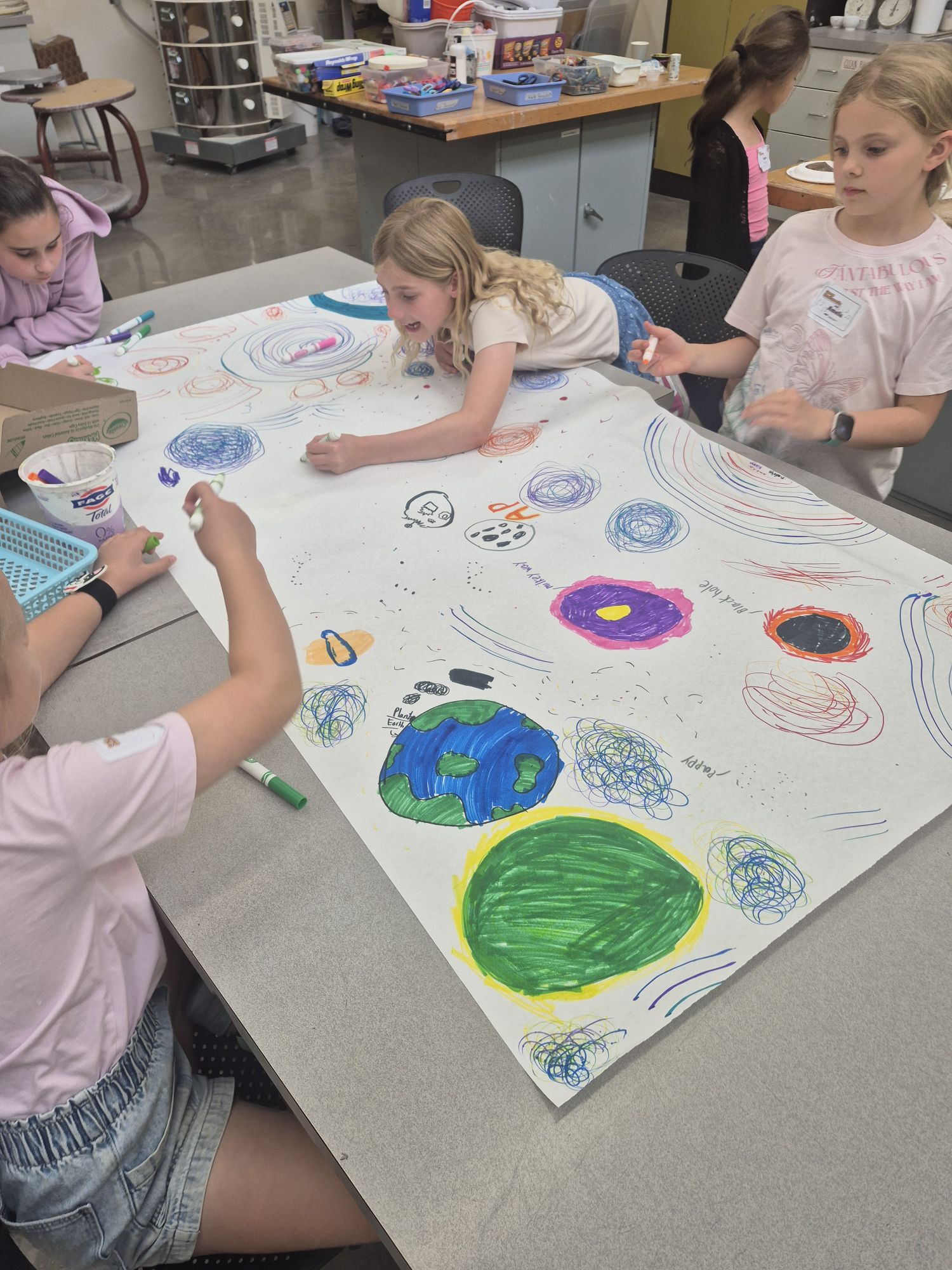 girls at a table drawing together on a large piece of paper