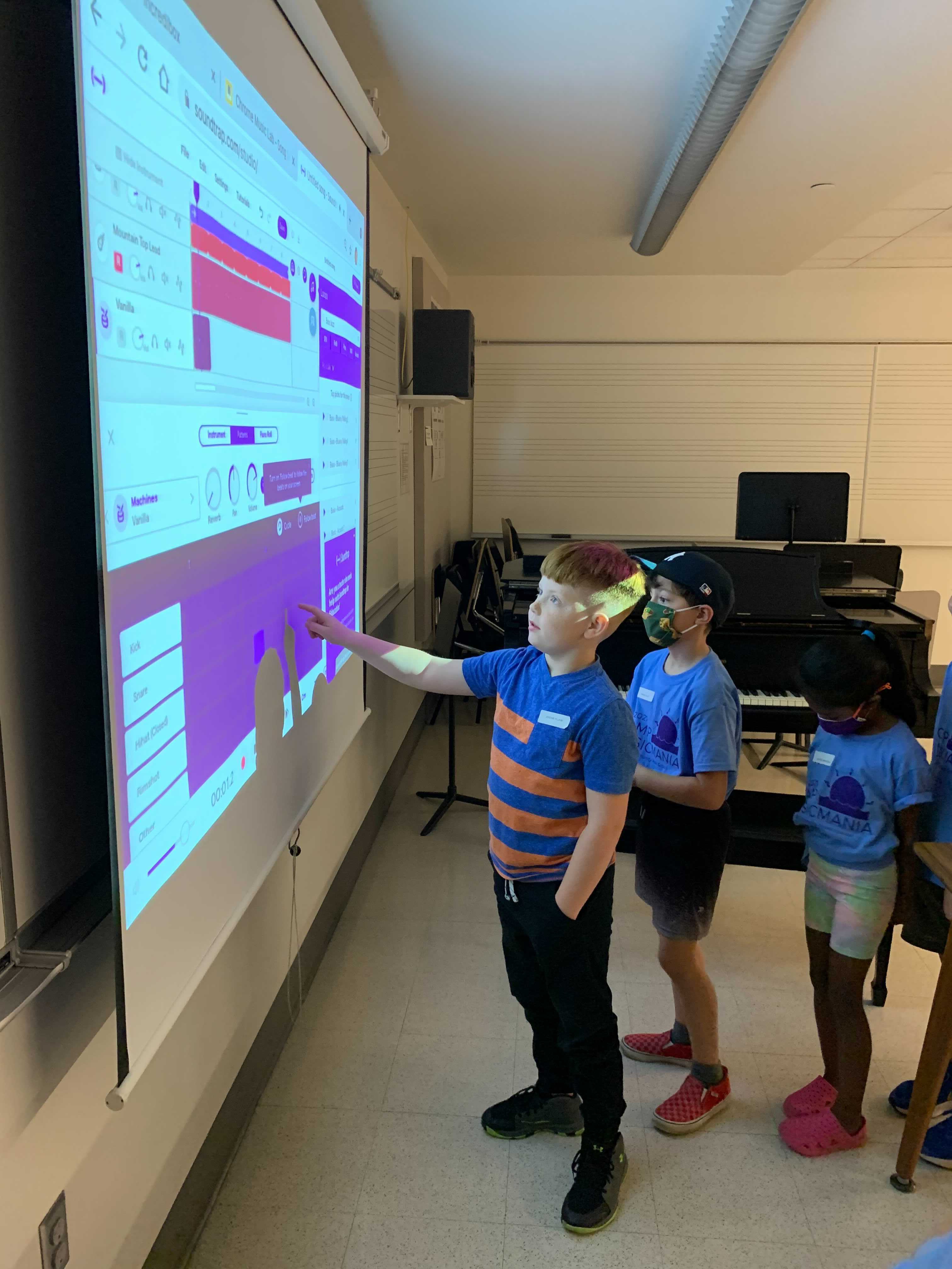 Three young students standing in front of a screen in a classroom, with one pointing at something on the screen.