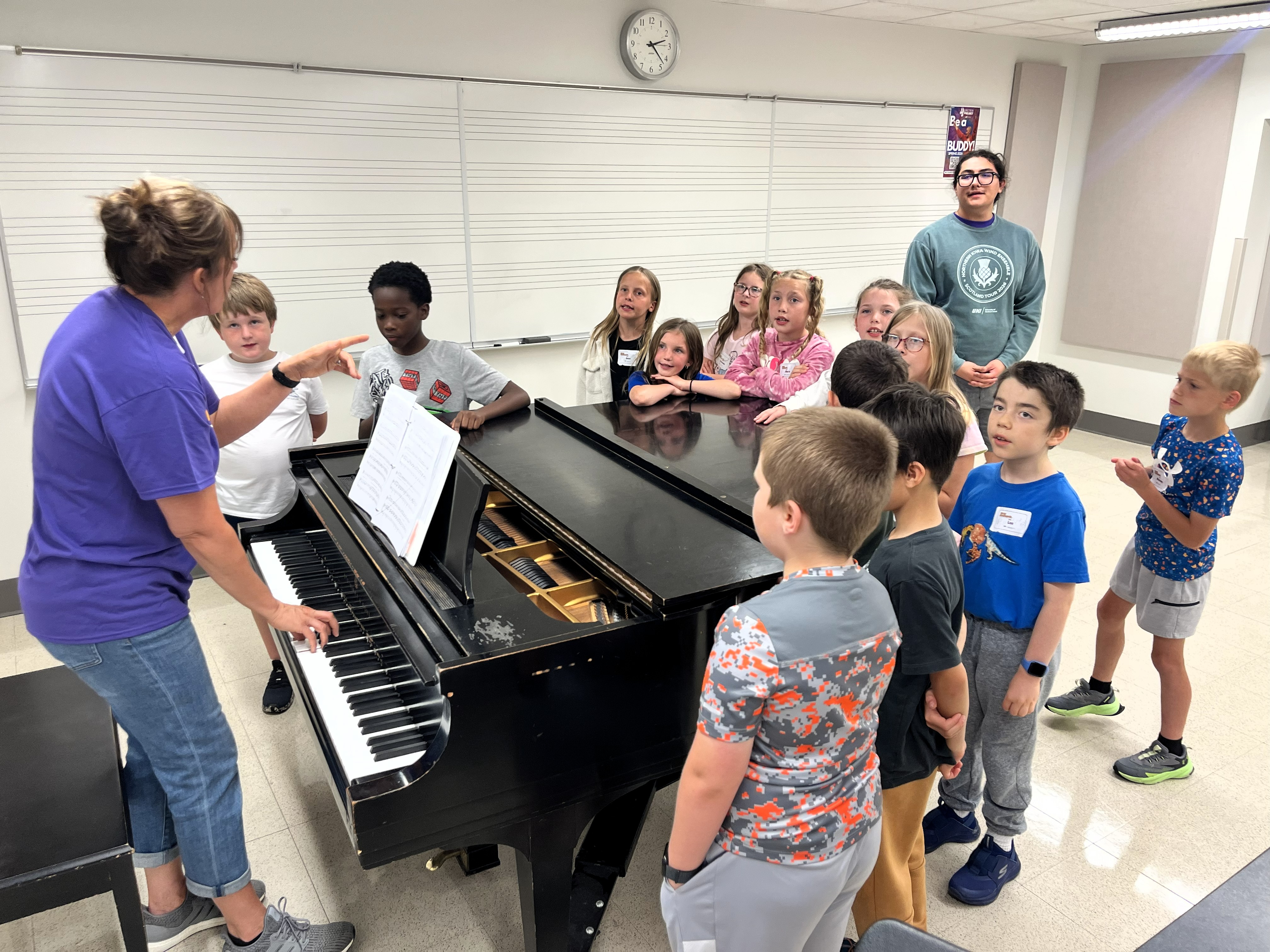 Kate Saulsbury teaching while standing and playing a grand piano. Kids surround the piano.