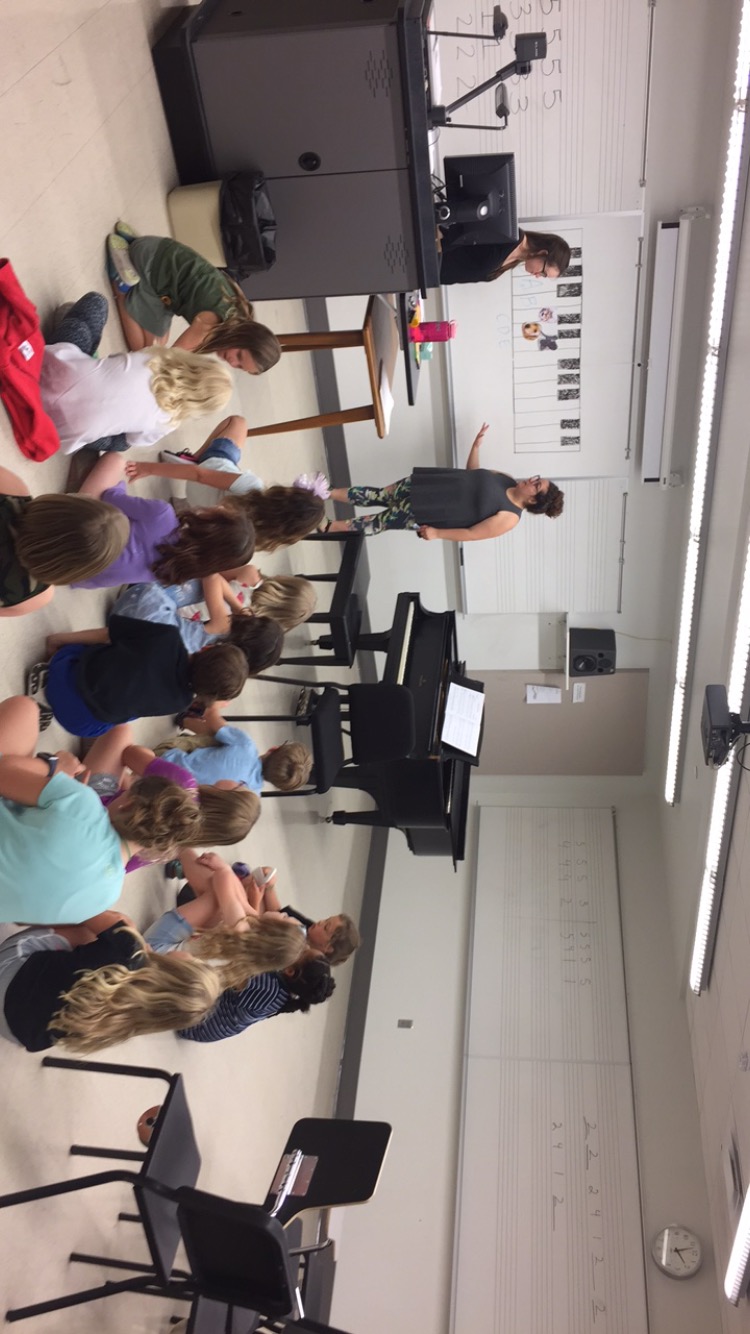 kids sitting on the floor in a classroom, watching teachers pointing at a drawn keyboard on a dry erase board on the wall.