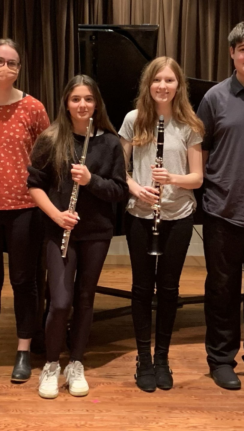 Girl on the left wearing black, standing and holding a flute next to another girl, wearing white and black, holding a clarinet in front of her.