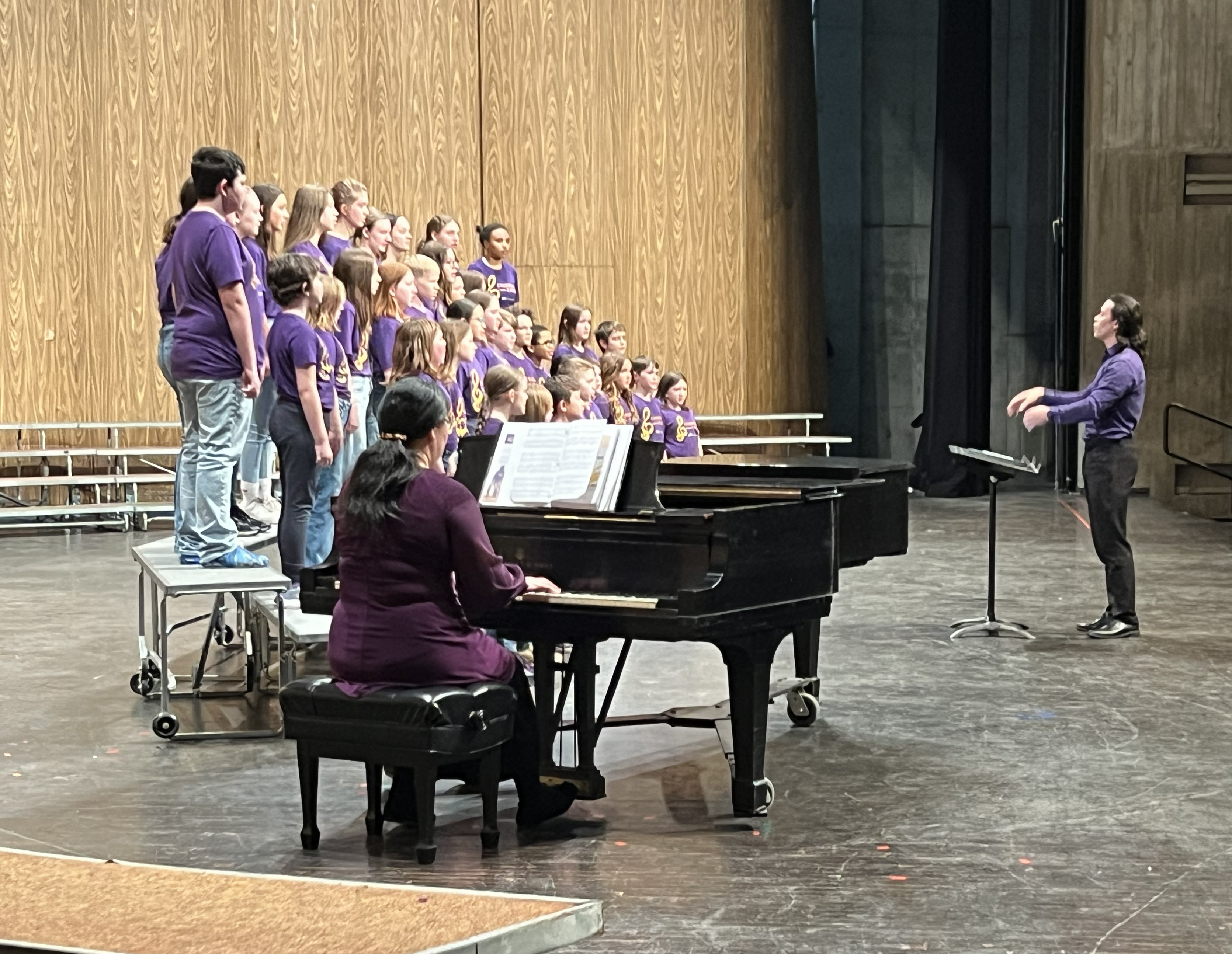 Looking from stage left, watching the Children's Choir performing. Pianist playing the piano, choir members standing on risers, and director in front of choir.