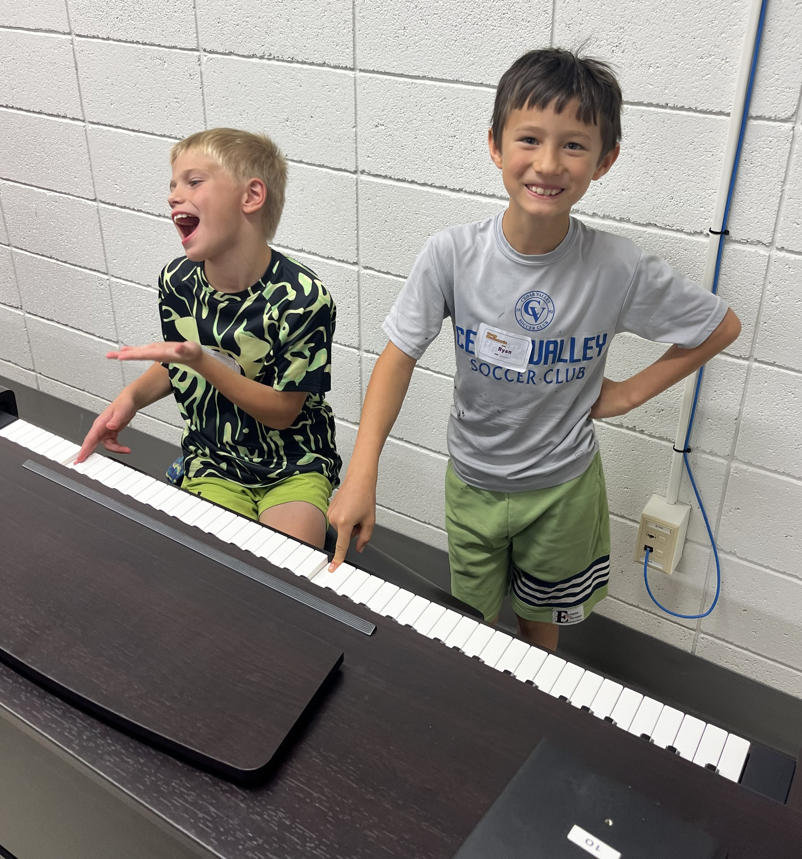 Two boys at a piano. One is sitting and laughing and the other is standing and smiling