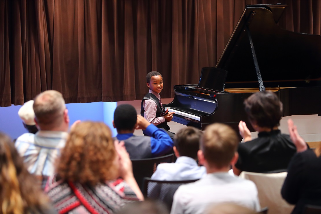 boy sitting at a grand piano smiling and facing the audience as the audience members applaud.